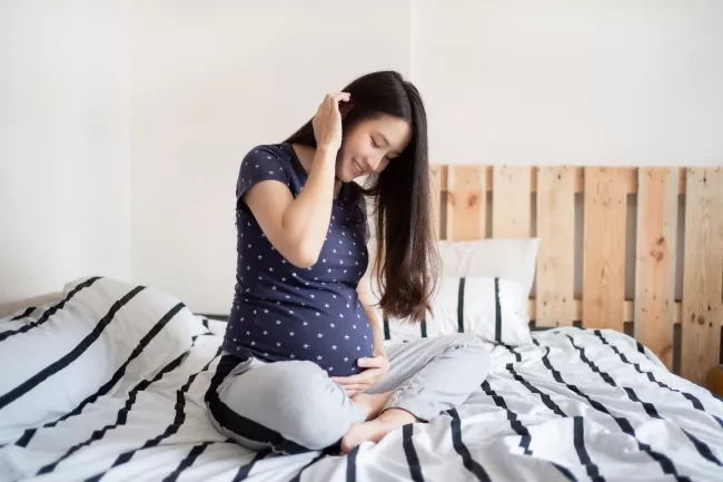 Pregnant mom sitting on her bed with a hand over her belly. 