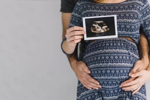 Soon to be father hugging his pregnant wife from the back and holding up an ultrasound photo